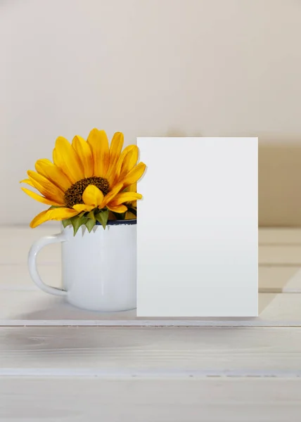 Greeting card mockup with sunflowers in a white cup. Empty postcard and enamel cup with space for text. White wooden background. Atmospheric moments.