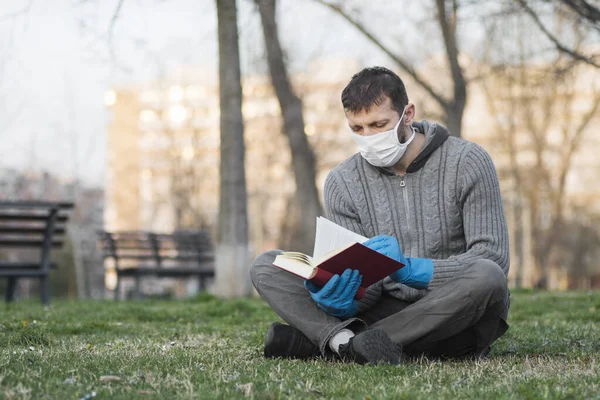 The man in protective mask and gloves is reading book and sitting on the grass outdoors.