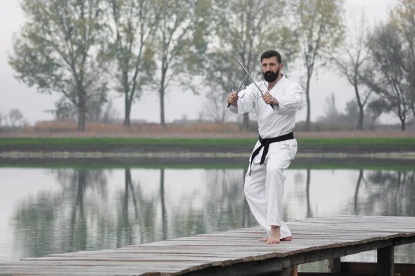 The man in white kimono is practicing martial arts with sai weapons outdoors on lakeshore in nature.