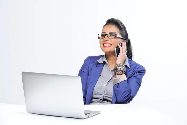 Beautiful young woman sitting at office desk and talking on cell phone isolated on white background — Stock Photo, Image