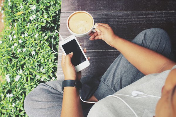 Женщина играет в смартфон с наушником и кофе в саду
