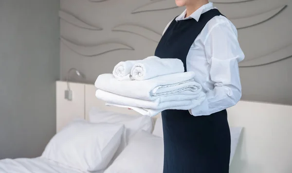 ホテルのルームサービスの客室を作る 若い女性の部屋メイドは 折り畳まれた白いバスタオルのスタックを保持し 客室内のベッドの横に立って — ストック写真