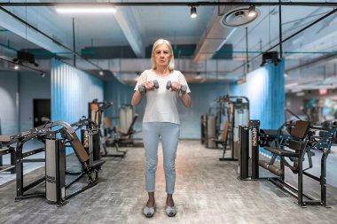 Kendine güvenen son sınıf öğrencisi bir kadın modern bir spor salonunda dambılları kaldırarak ağırlık kaldırıyor. Sahnede fitness, güçlendirme, sağlık ve istikrarlı ilerleme vurgulanıyor.