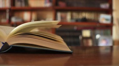 Kapağı açık bir kitap, arka planda bulanık kütüphane rafları olan sıcak ahşap bir masanın üzerinde duruyor. Sakin bir okuma alanında çalışma, bilgi ve hayal gücünü öneriyor..