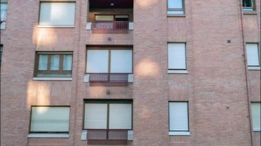 Birden fazla pencere ve küçük balkonların yer aldığı tuğladan bir apartman cephesi, dokulu tuğla duvarlarda sıcak güneş ışığıyla kentsel yaşam ve konut mimarisini vurguluyor.