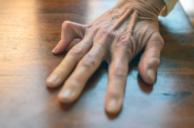 Ahşap bir masada dinlenen yaşlı bir elin yakın çekimi. Derin kırışıklıklar, doku ve tetik parmağı..