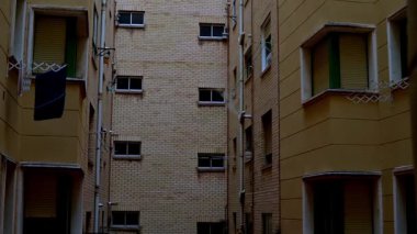 Eğil. Pencerelerin arasında kirli çamaşırların asılı olduğu sarı tuğlalı binalarla çevrili sessiz bir sokak. Sahne günlük şehir hayatını, yerel ritmi ve paylaşılan mekanın mahrem detaylarını yakalıyor..