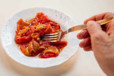 Beyaz seramik tabakta zeytinyağıyla korunmuş ızgara kırmızı biber yiyen bir el, arka planda hafif bir şekilde servis edilen ev yapımı basit bir vejetaryen yemeğini gösteriyor.