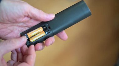 Bir insanın elleri iki tane AA pil yerleştirmek için parlak siyah bir cihaz açar. Basit, taşınabilir bir güç kaynağı. Uzaktan kumandalar, aletler ve günlük elektronik aletler için tipik..