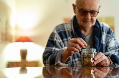 Gözlük takan ve ekose bornoz giyen yaşlı bir adam, tasarruf, bütçe ve emekliliği gösteren cam bir kavanoza bozuk para koyar. Yumuşak ışıklandırmalı ve yansıtıcı masa yüzeyli sıcak, samimi bir sahne..