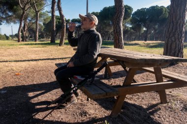 Manzaralı bir ormanda piknik bankında oturmuş su içen bir adam. Yürüyüş ekipmanları ve parlak güneş ışığı huzurlu ve ferahlatıcı açık hava atmosferini arttırır..