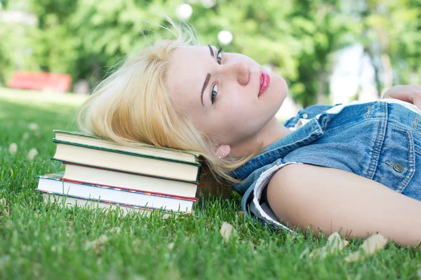 Hermosa joven sonriente tumbada en la hierba y leyendo libro azul, parque verde de verano. Mujer estudiante afuera en el parque. Feliz joven estudiante universitario de etnia mixta europea y caucásica . — Foto de Stock