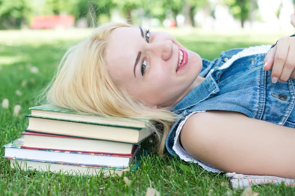 Beautiful smiling  young woman lying on grass and reading blue book, summer green park. Female student girl outside in park. Happy young university student of mixed European and Caucasian ethnicity.