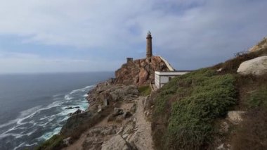 Aerial establishing shot of a historic lighthouse on Costa da Morte cliff overlooking the Atlantic, with dramatic waves crashing against the rugged coastline.