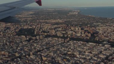 Plane-window view: red-and-white wingtip over a dense coastal city, showing shoreline and long shadows in warm low sunlight during approach.