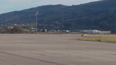 Dağlı geniş bir pistte sarı bakım aracı taksileri ve sakin gündüz havaalanı operasyonları sırasında arka planda kontrol kuleleri.