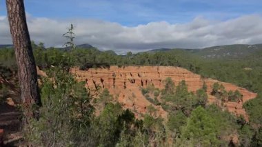 Sahne: Kırmızı toprak kayalığı, çam gövdesi ve derin mavi bir gökyüzünün altındaki orman dağları yavaş bulutlar, parlak güneş, kıtır gölgeler ve sakin.