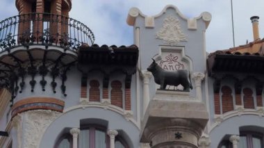 Yavaş bir tava, İspanya 'nın Teruel şehrinde koyu renkli bir boğa heykeli, işlenmiş demir balkon, yumuşak, bulutlu ışık altında tuğla ve mavi aksanlı süslü bir Art Nouveau cephesini gözler önüne seriyor..