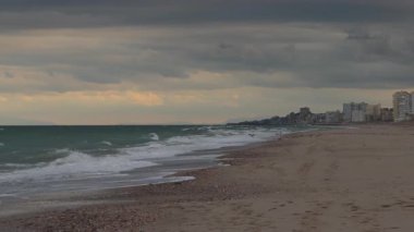 Güçlü yeşil dalgalar, günbatımında kumlu bir sahile çarpar dramatik fırtına bulutlarının altında, ufuktaki uzak sahil şehri ufkuyla..