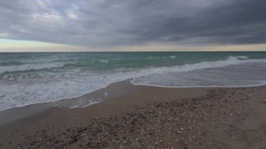 Kasvetli okyanus dalgaları bulutlu gündoğumu / günbatımı altında karanlık bir çakıl taşının üzerinde süzülüyor, hipnotik, sakin bir deniz manzarası yaratıyor..