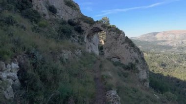 Dar bir dağ yolunda yavaşça ilerler doğal bir taş kemere doğru, kayalık uçurumları, yeşil bir vadiyi ve yumuşak akşam ışığında uzak tepeleri ortaya çıkarır..