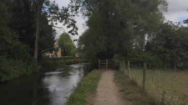 Yeşil bir yolda huzurlu nehir kenarında yürüyüş, hafif bir esinti, bereketli bir manzara, ve arka planda klasik İngiliz taş evleri.