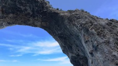 Yavaş bir tava, dokulu taş, yeşil tepeler ve sakin bir Akdeniz manzarasında mavi gökyüzü ile muazzam bir kaya kemerini ortaya çıkarır..