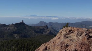 Pico de las Nieves 'ten statik panoramik manzara: kayalık ön plan, çam kaplı dağlar, bulutlar denizi ve açık mavi gökyüzü altında uzak bir ada.