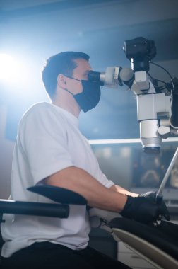Mikroskopta oturan dişçi hassas klinik prosedürü, odaklanmış duruşu ve eldivenli elleri gerçekleştiriyor.,