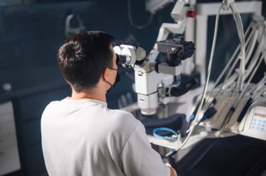 Mikroskopta oturan dişçi hassas klinik prosedürü, odaklanmış duruşu ve eldivenli elleri gerçekleştiriyor.,