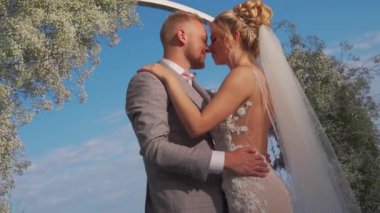 The bride and groom kiss against the background of the arch and the blue sky. Close up