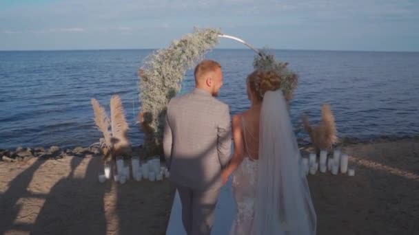 The bride and groom go to arch and kiss against the sea — Stockvideo