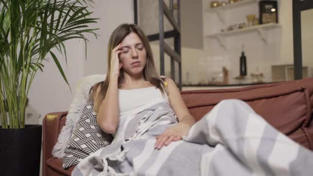 Die junge Frau hat Kopfschmerzen. Sie liegt auf dem Sofa und schaut auf das Thermometer — Stockvideo