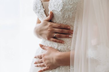 Gri arka planda güzel bir gelin. Kadının üzerinde uzun, dantelli ve arkası açık bir gelinlik var. Ellerinde buket.