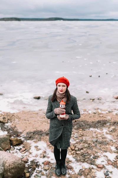 Winter portrait of young woman in a coat and red hat holding decorative lighthouse and standing at the shore of frozen sea. winter, travel, sea background. windy weather, ice seaside