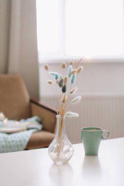 Bir fincan kahve ve beyaz arka planda kurumuş çiçekli şeffaf bir vazo.