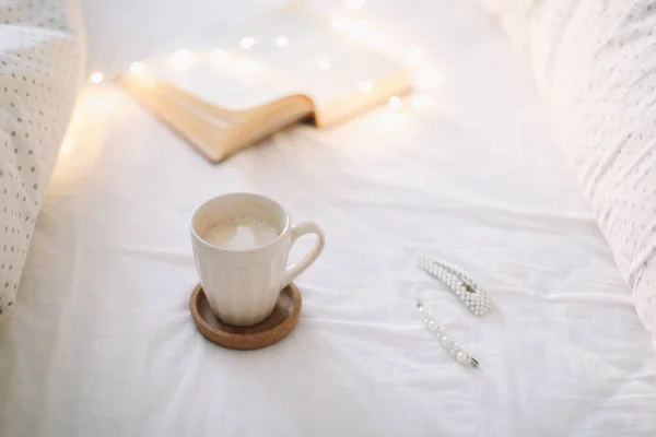 Taza de café con espuma de leche y un libro sobre una sábana blanca. — Foto de Stock