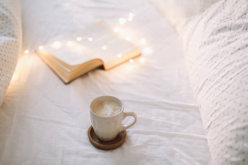 Coffee cup with milk foam and a book on a white bedsheet. 