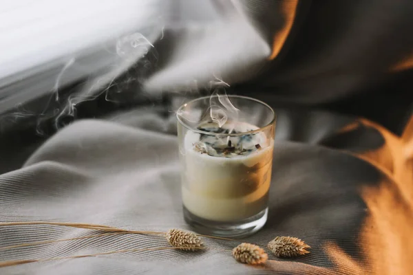 Aromatic scented candle with smoke and dried flowers on beige textile background. — Stock Photo, Image