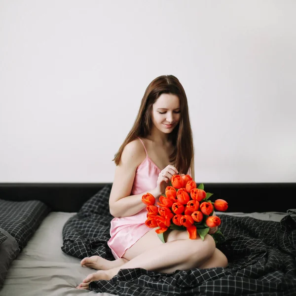 Joven hermosa mujer sentada en la cama y sosteniendo un ramo de tulipanes rojos — Foto de Stock