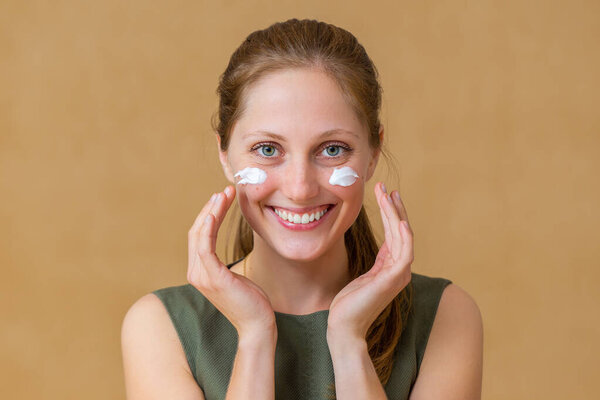 Happy woman applying white cream on her face and looking into camera.