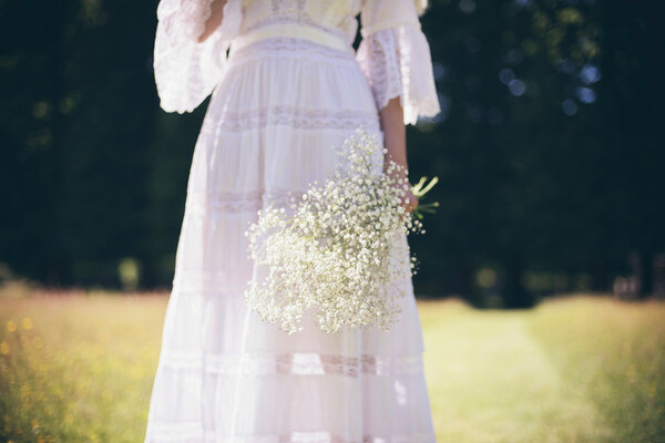 Partial view of young bride in white dress holding white flowers standing in meadow at sunny summer day