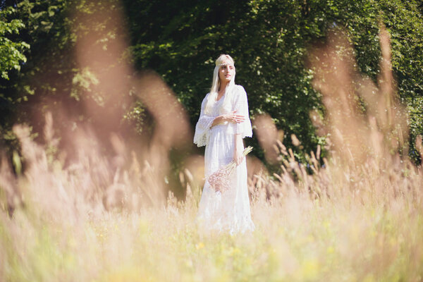 Beautiful blonde hippie bride in white dress holding bouquet standing in high grass at sunny summer day