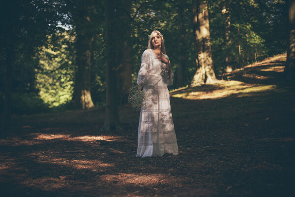 Beautiful blonde bride in white dress holding white flowers standing in dappled sunlight of forest