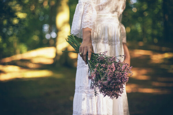 Close-up hand of bride in white dress holding purple flowers standing in forest at sunny day