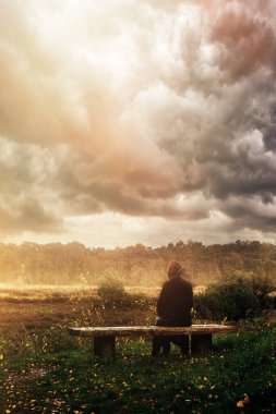 Woman in black coat sits on wooden bench in nature under cloudy sky. clipart