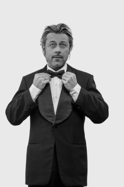 Smokinli bir adam beyaz duvarın önünde papyon tutuyor. Siyah beyaz fotoğraf.