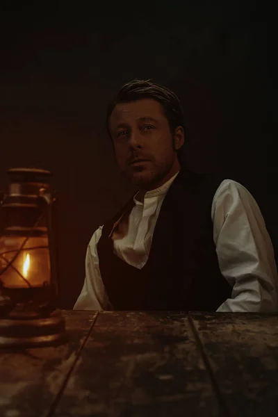 Vintage victorian style man behind wooden table with historical oil lantern.