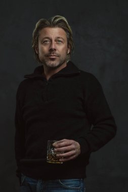 Siyah yün kazaklı mutlu sarışın adam elinde bir bardak viski tutuyor..