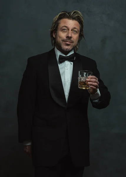 Satisfied blonde man with stubble beard in tuxedo holding glass of whisky.